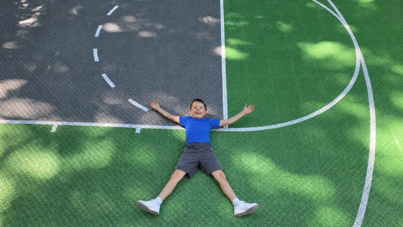 Child lying with arms outstretched on a finished green and charcoal modular sports court with white game lines.